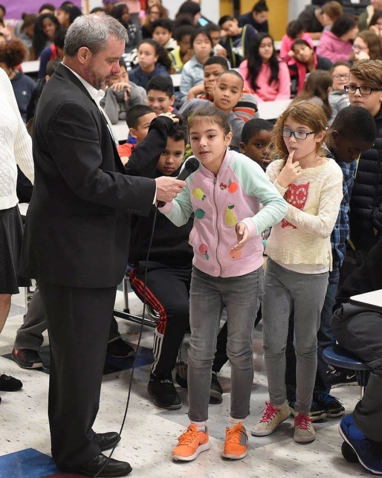 A man holding a microphone for a child at the front of a line of other children