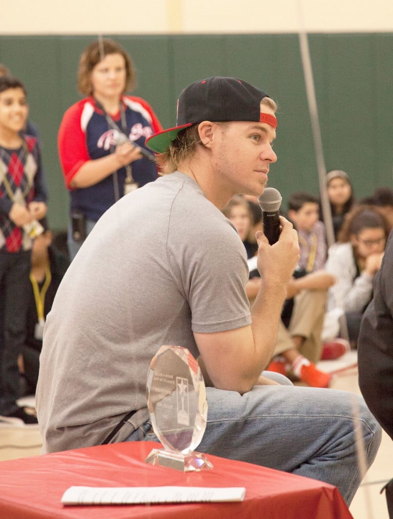 Brock Holt seated on a stool, talking with a microphone, next to a table with a small trophy with children seated in the background