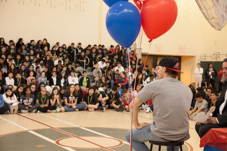 Brock Holt addressing a large group of children seated on the floor and in the stands of a gymnasium