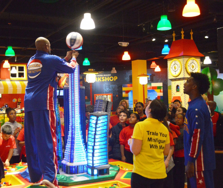 A Harlem Globetrotter spinning a basketball on his figure in front of another Globetrotter, a woman, and a group of children