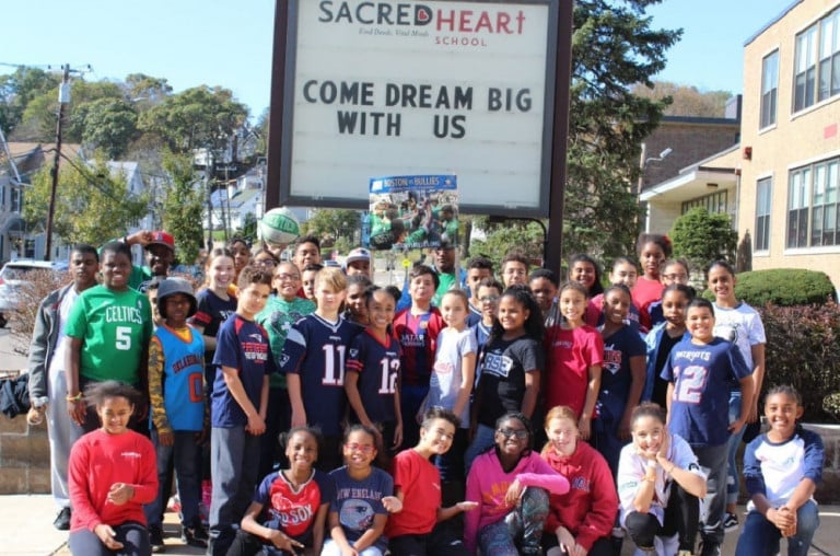 A group of children posing in front of a Sacred Heart school sign that says "Come Dream Big With Us"