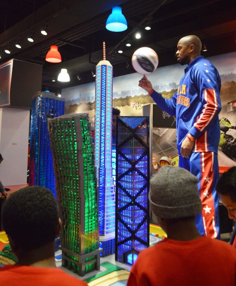 A Harlem Globetrotter spinning a basketball on his finger next to a four lego skyscrapers and in front of a group of children