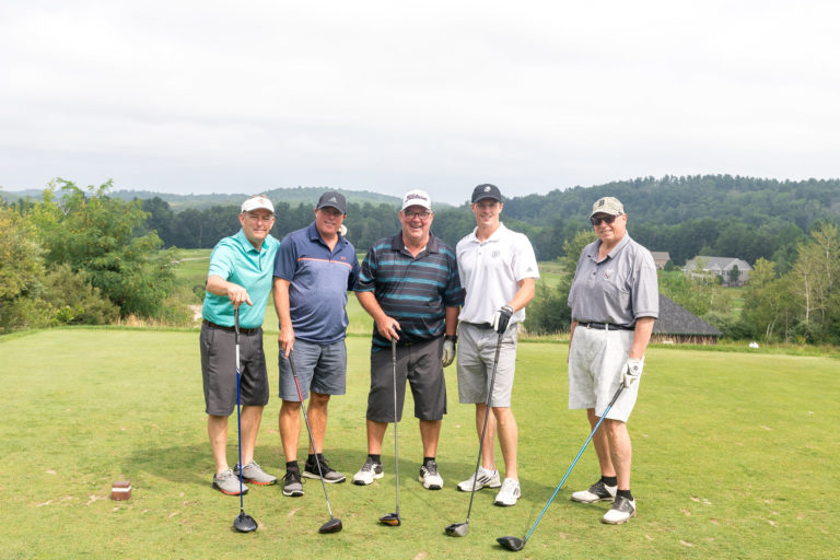 Group of five men smiling standing next to each other and smiling at a golf course.
