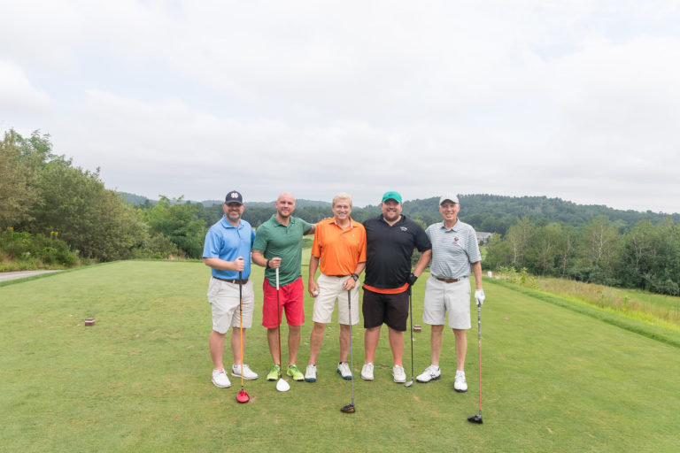 Five men are holding their golf clubs, standing next to each other, and smiling at the camera.